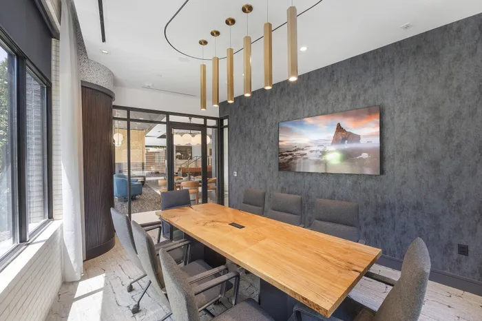 Modern conference room with large exterior and interior windows, a flat-screen TV, and 8 upholstered office chairs around a wood conference table.