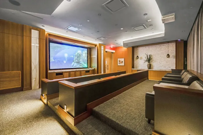 Theater with three rows of large, leather seats and a third row of lecture-hall-style rolling chairs behind a long desk. There is a projector screen on the wall and blackout curtains around the windows.