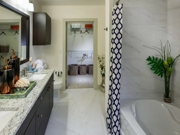 Bathroom with a walk-in closet, dual vanity with a gray granite countertop, white marble-style floors, and full-size soaking tub.