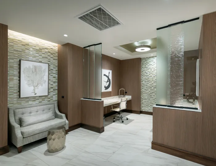 Two desks with rolling leather office chairs are tucked behind textured glass dividers. The room has a loveseat in the foreground, white marble floors, and wood-paneled walls with mosaic insets.