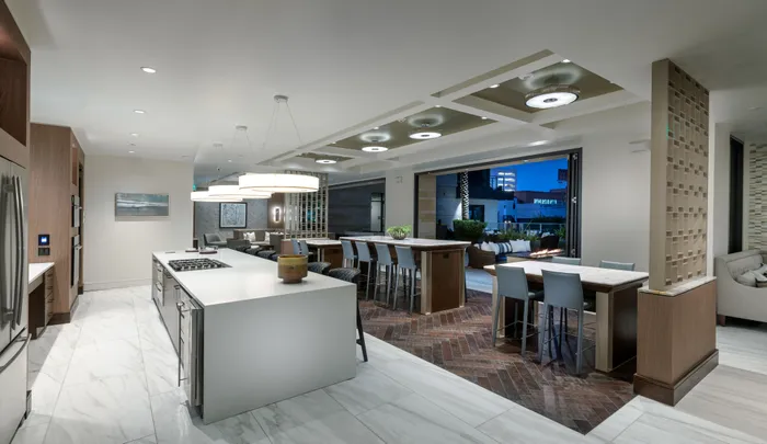 Clubhouse dining area including a catering kitchen with two ovens, a gas stove on a large island with 7 barstools, and 3 dining tables accommodating 4 to 8 chairs. The large floor-to-ceiling windows have been slid back to allow access to an outdoor seating area with a firepit.