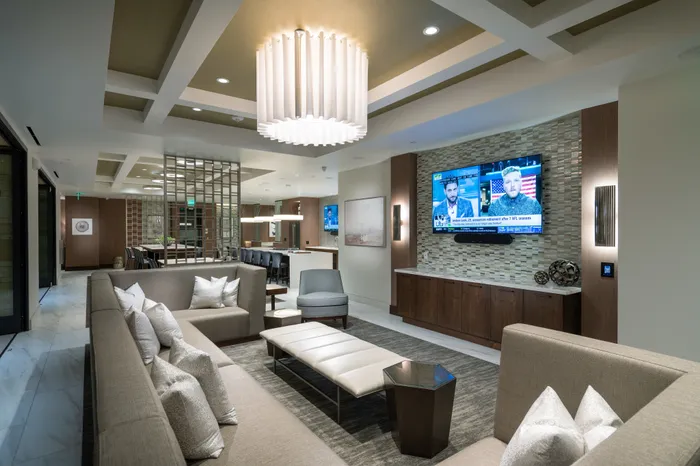 A seating area adjacent to the clubhouse kitchen and dining area with a large wraparound sectional couch in front of an HDTV.