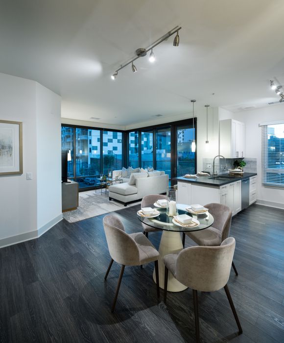 Open-concept kitchen, dining, and living space with dark wood-style floors, white cabinets, a kitchen peninsula with dark granite countertops, and two full-width floor-to-ceiling windows in the spacious living room.