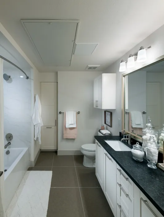 Spacious bathroom with warm gray tile floors, a large soaking tub with a shower curtain, white cabinets, black quartz countertops, and a large framed mirror.