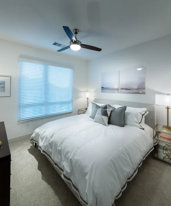 Bedroom with a king-sized bed, two side tables, a dresser, a large window, white walls, a ceiling fan, and beige carpeting.