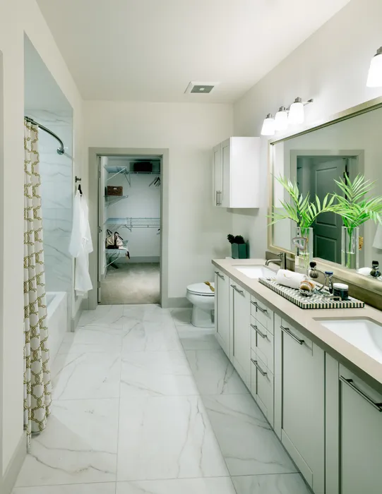 A bright bathroom with a dual vanity, large mirror, white cabinets, a large tub, marble tile on the floor and bathtub walls, and a walk-in closet.