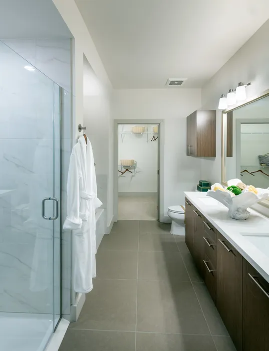 Bathroom featuring a frameless glass door walk-in shower, a dual vanity with dark wood cabinets and a white quartz countertop, and a walk-in closet.