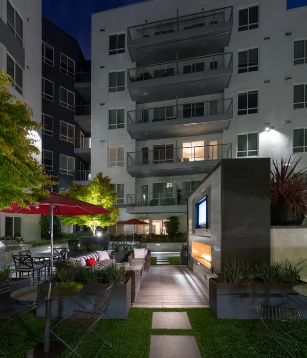 Outdoor seaing area including several patio tables and chairs with red umbrellas, a large sectional couch, and an HDTV mounted over a modern fireplace in a marble facade.