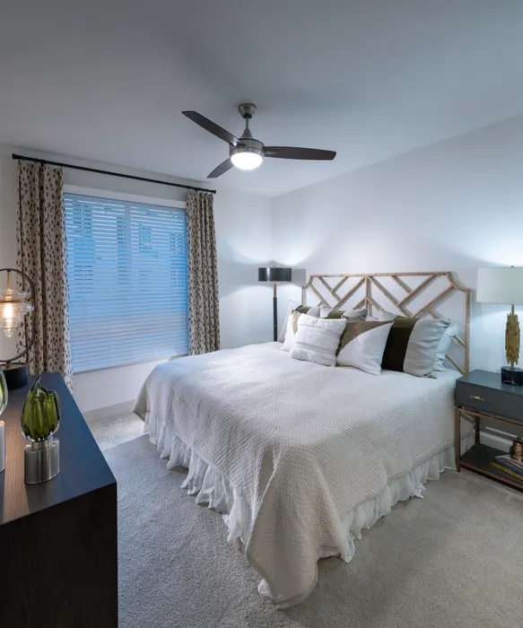 Bedroom with king-sized bed, side table, dresser, large window, ceiling fan, beige carpet, and white walls.