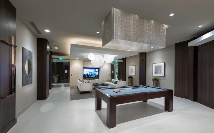 A clubhouse with dark wood-paneled walls, a billiards table under a rectangular chandelier with hanging silver chains, and two long white couches in front of a large HDTV.