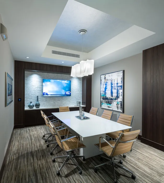 A 8-person conference table with rolling leather chairs in a room with gray and wood-paneled walls, neutral carpeting, abstract art, and an HD presentation TV.