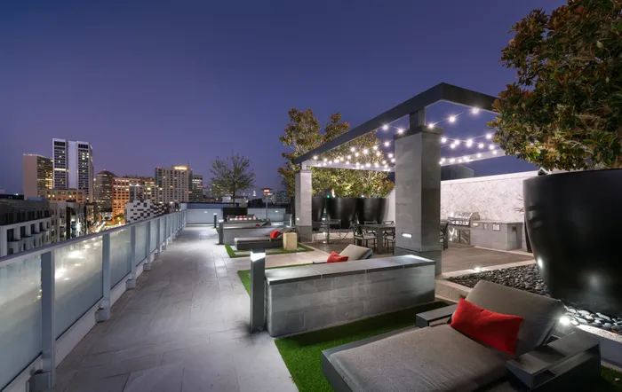 A landscaped rooftop entertaining area with a dining table, grilling station, and several lounge chairs facing a city skyline view.