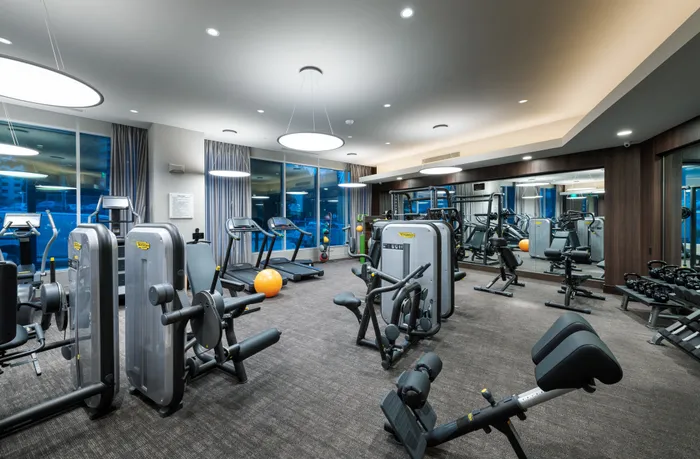 Indoor gym with large windows, carpet floors, treadmills, ellipticals, weight machines, kettlebells, and two mirror walls.