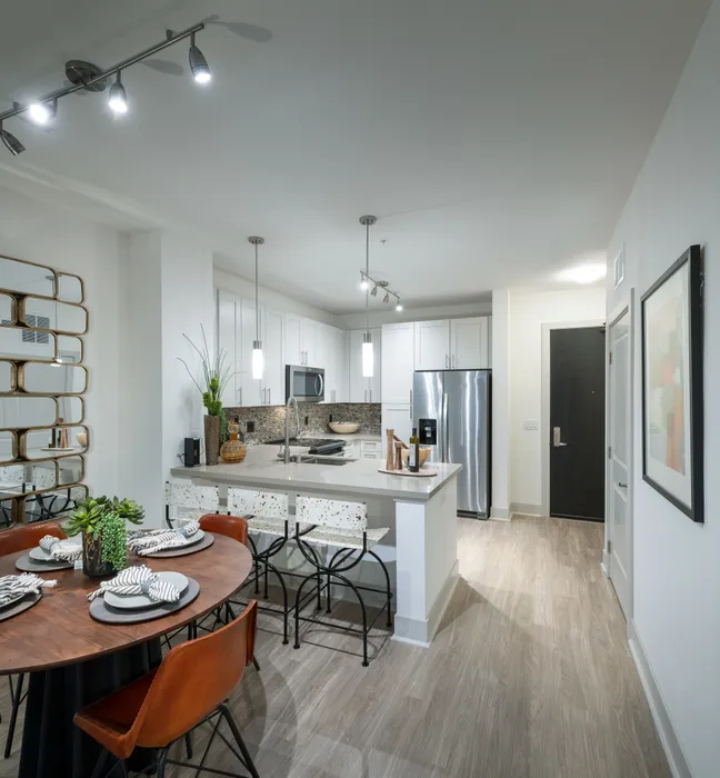 Open-concept kitchen and dining area with white walls, light wood-style floors, a four-person dining table, three barstools at the kitchen peninsula, white cabinets, and stainless steel appliances.
