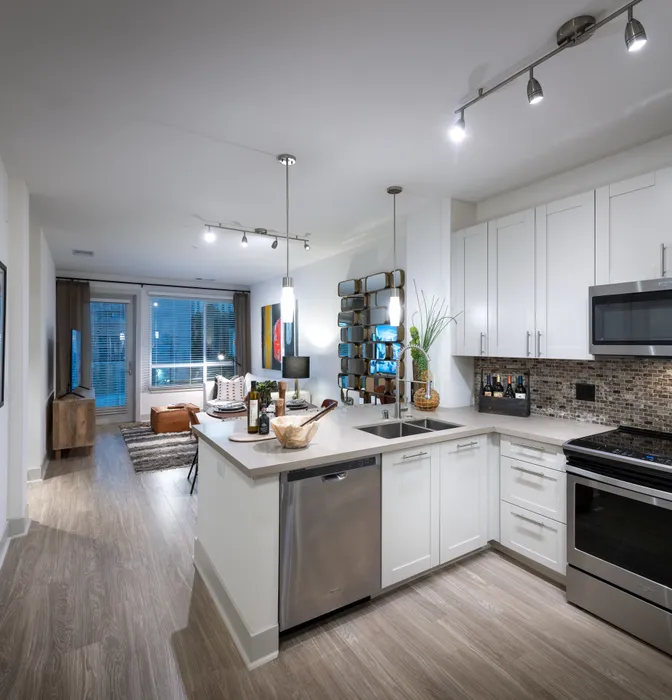 Open-concept kitchen, dining, and living space with white walls, light wood-style floors, white cabinets, stainless steel appliances, and a large window and glass door leading from the living area to the balcony.