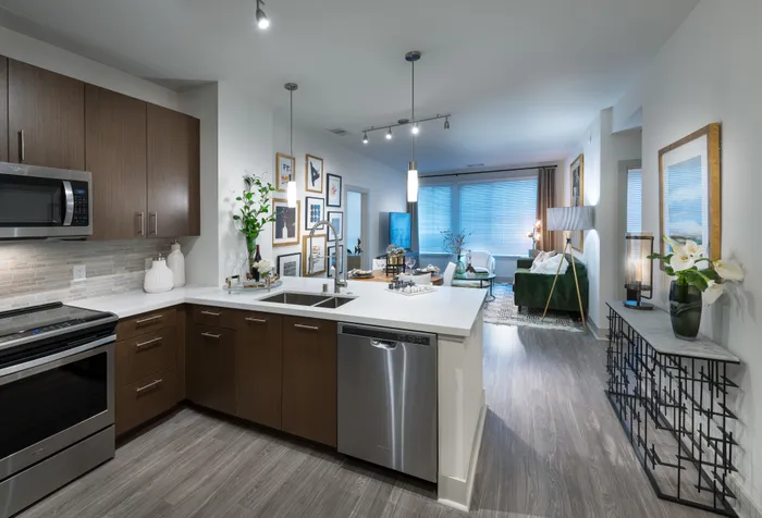 Open-concept kitchen, dining, and living space with ash-gray wood-style floors, dark modern cabinets, stainless steel appliances, white quartz countertops, and a spacious living area with a large window.