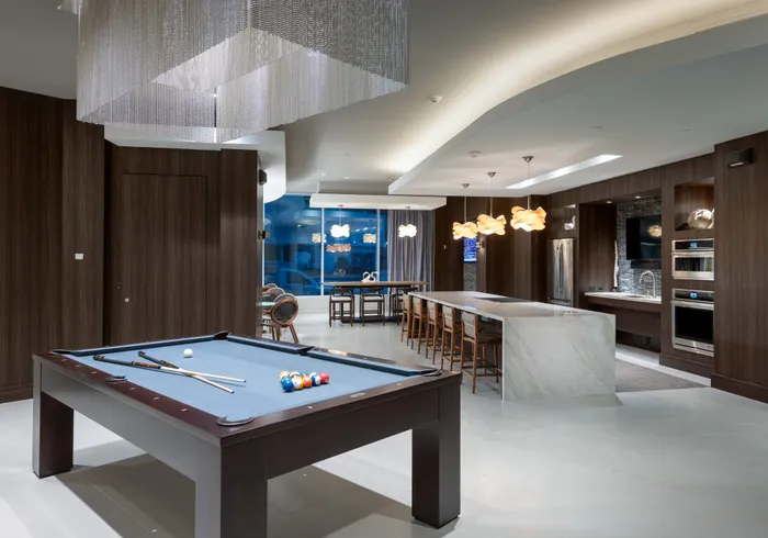 A clubhouse with dark wood-paneled walls, a billiards table under a rectangular chandelier with hanging silver chains, and a resident catering kitchen with two ovens, a large marble island with barstools, and an HDTV.