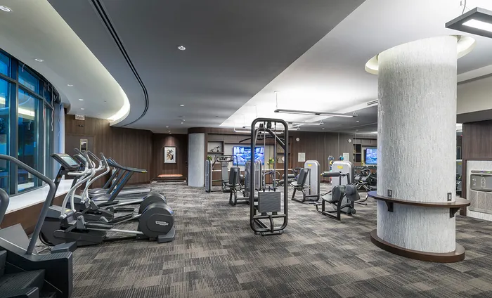 Alternate view of the gym with weight machines, an HDTV mounted on the wall, a full-width mirror wall, carpeted floors, and a bank of treadmills, ellipticals, and stair climbers in front of a large, curved window.