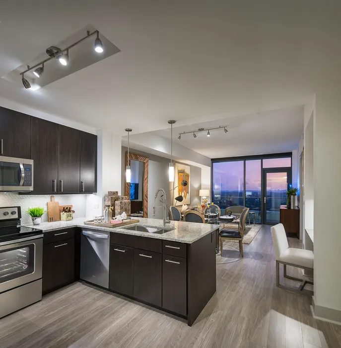 The same open-concept apartment with a pink, purple, and orange city sunset view through the full-width floor-to-ceiling window. The space features ash-gray wood-style floors, dark modern cabinets, stainless steel appliances, granite countertops, and a spacious living area.