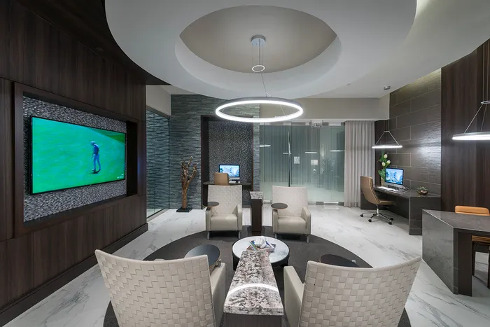 A modern work space with two Mac computers at separate marble desks, a sitting area with 4 chairs in front of an HDTV, and a ring-shaped light fixture mirroring a large circular suspended ceiling detail.