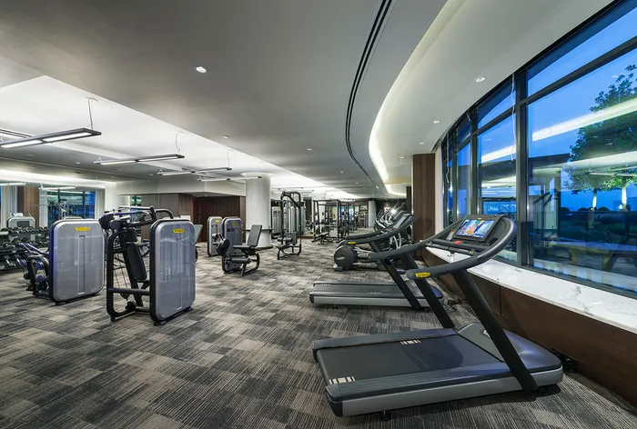 Extensive indoor gym with weight machines, dumbbells, a full-width mirror, carpeted floor, and treadmills in front of a large, curved window.