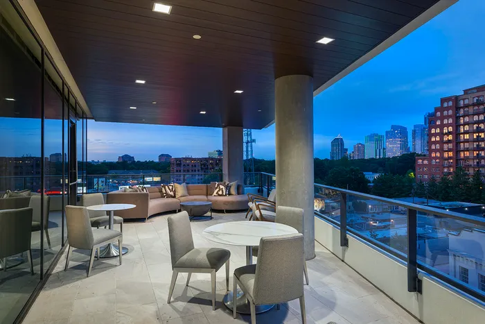 An open-air loggia with a view of the city skyline at dusk, a wraparound glass railing, a seating area with a curved couch and armchairs, and multiple sets of white tables and chairs.