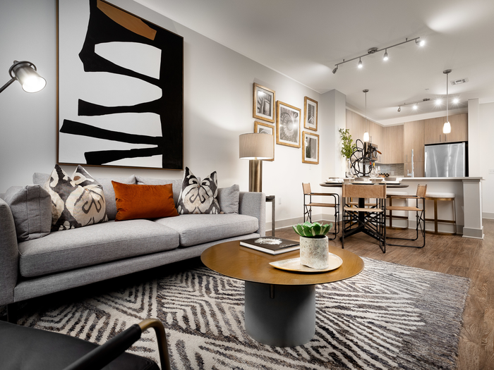 Open-concept living room, dining area, and kitchen with a light gray couch, small coffee table, abstract art on the wall, a round dining table with 4 chairs and a kitchen island with barstools.