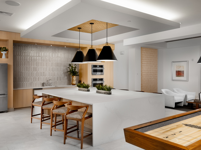 Clubhouse with white marble floors, a shuffleboard, a light wood-paneled wall next to stainless steel appliances, a large white marble island with 4 brown barstools and modern artwork and decorations.