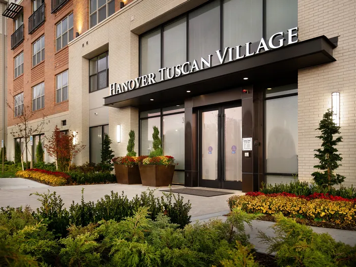 Entrance to Hanover Tuscan Village apartment building with an orange and cream exterior, glass doors to the entrace, windows on each level and a sidewalk lined with plants and red and yellow flowers.