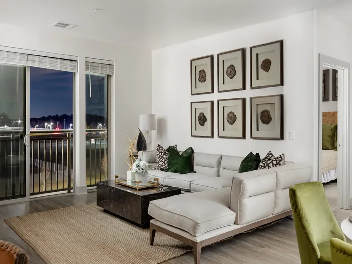 Living room with a white sectional couch, brown marble coffee table, artwork on the walls, large windows opening to a balcony and a hallway and door leading to another room.