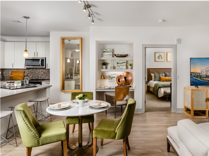 Rendering of an open-concept dining area, with a round white quartz table, 3 upholstered green chairs, a built-in desk station with shelves and and opening to a bedroom.