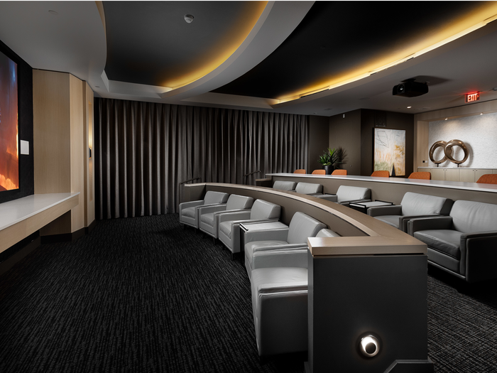 Theater with two rows of large, leather seats and a third row of lecture-hall-style chairs behind a long desk. There is a projector on the ceiling and blackout curtains on the wall.