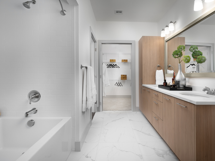 Spacious bathroom with white marble floors, a large bathtub with a showerhead, light wood cabinetry and a dual vanity. There is an entrance to a walk-in closet.