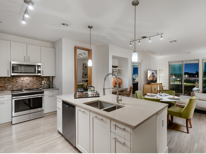 Open-concept kitchen, dining, and living room area with white cabinetry, stainless steel appliances, light wood-style floors, modern light fixtures, a dining table with 3 green chairs and large windows leading to a balcony.