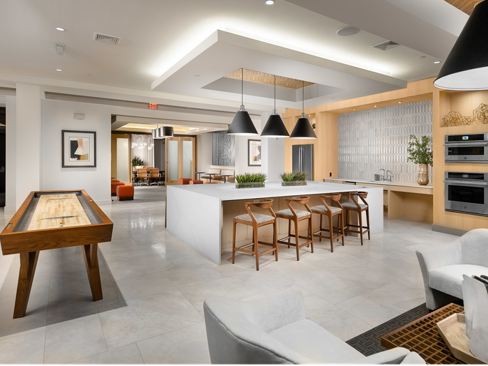View of the clubhouse from a different angle, with 2 white chairs, a coffee table, a shuffleboard, and an open entrance to another indoor lounge.