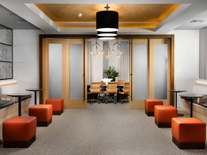 Meeting area with booth-style seats, small round black tables and 6 red square stools. There is an entrance to a private meeting room with fully opaque glass and wood framed sliding doors.