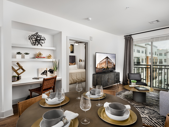 Dining area with a round wooden table, chairs and a built-in work desk with 2 shelves decorated with modern art. The room opens to a living room with an HDTV and large windows.