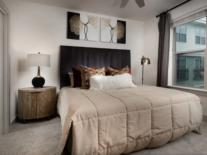 Spacious bedroom with a side table and lamp, a standing lamp, modern artwork and a window with curtains.