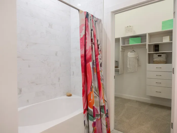 Bathroom with an oval soaking tub, floral shower curtain, and marble tiling to the ceiling. A door leads into a large walk-in closet with beige carpet.