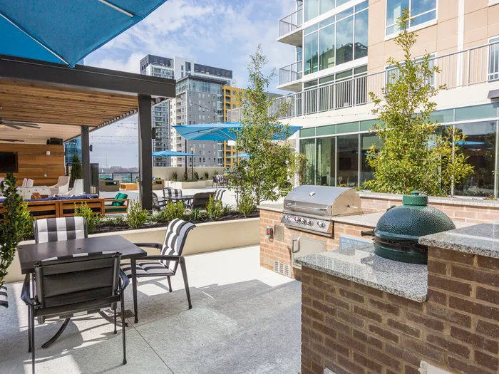 An outdoor grilling and dining area with a four-seat patio table, traditional stainless steel grill, and a Big Green Egg charcoal and ceramic grill. The prep spaces have granite countertops.