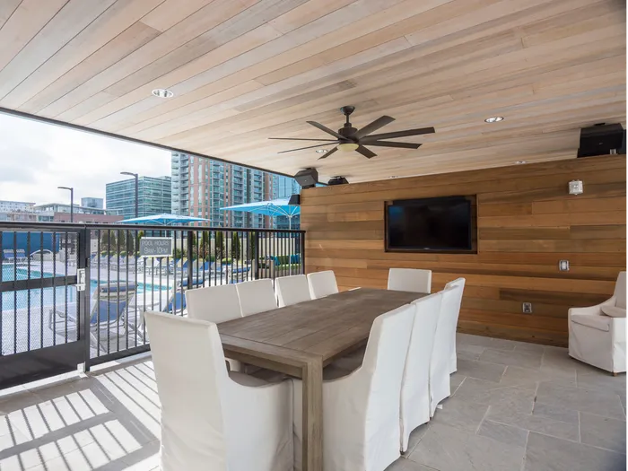 An open-air dining room by the pool enclosed with a gate, with a wood-paneled wall and ceiling, a flat-screen TV mounted on the wall, ten white upholstered chairs, and a ceiling fan.