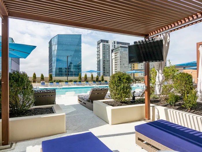 A wooden cabana space with two upholstered lounge chairs overlooking the pool and city skyline, enclosed by a raised landscaping bed with bushes.