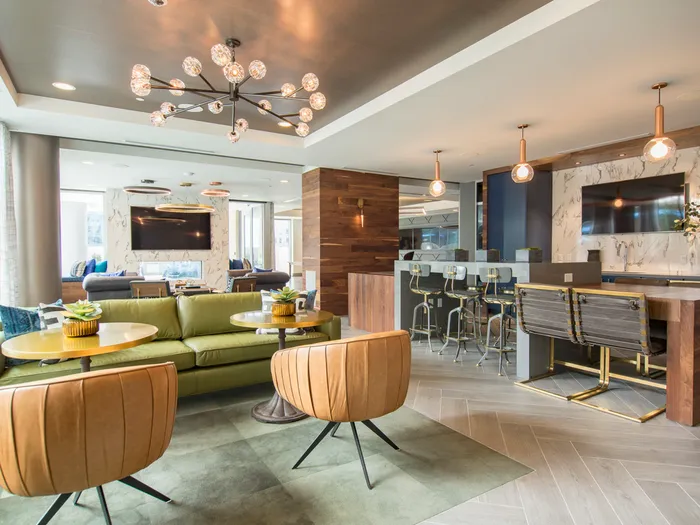 A bright lounge space with large windows, colorful leather couches and armchairs, a bar with an HDTV, a see-thru fireplace, wood-paneled walls, and modern light fixtures.
