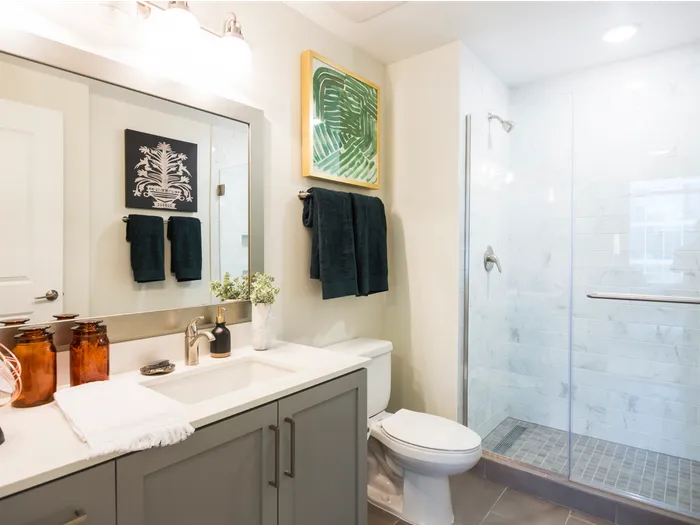 Bathroom with a glass-walled walk-in shower with floor-to-ceiling marble tile, a white quartz countertop with gray cabinets, and a large framed mirror.