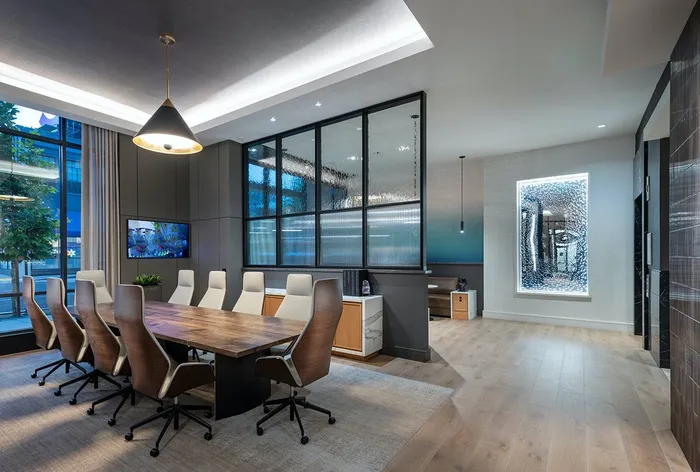 A modern conference room with a wooden 10-person table, midcentury-style office chairs, floor-to-ceiling windows, and a glass divider separating a seating area from the conference space
