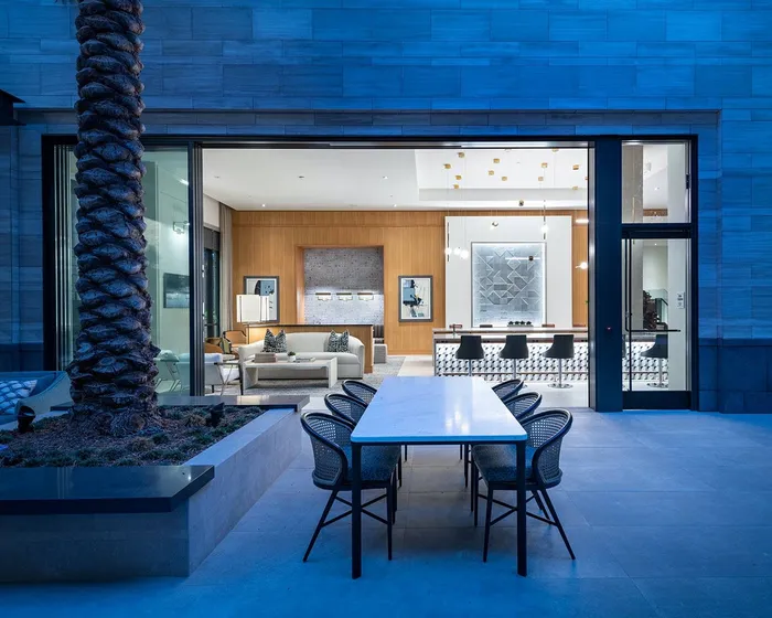An outdoor 6-person dining table with a large palm tree adjacent to the indoor clubhouse's open entrance