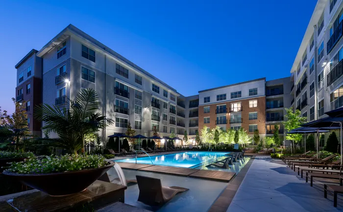 Image of a pool courtyard with brown and white apartment homes, a pool with lounge chairs in the shallow ends and surrounding area, landscaped areas with trees and an elevated deck with a table and chairs.