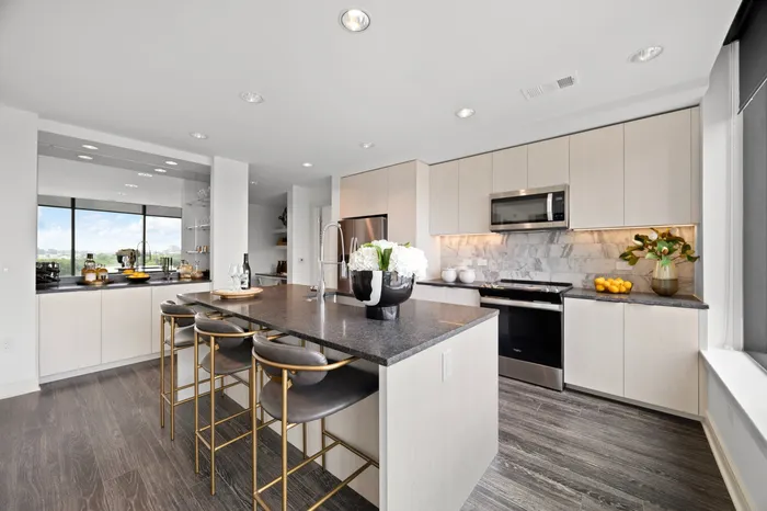 apartment kitchen with center island, built-in dry bar with mirror, light white wood cabinets, black stone counters, and brown wood-style flooring.