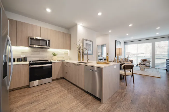 apartment kitchen with light wood cabinets, stone counters and backsplash, with dining and living area behind