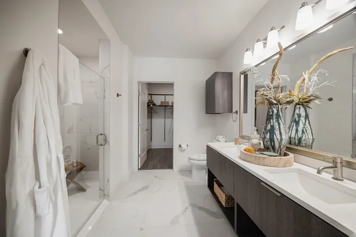 bathroom with dual vanity, framed mirror, glass frameless shower, white marble tile, and door to closet